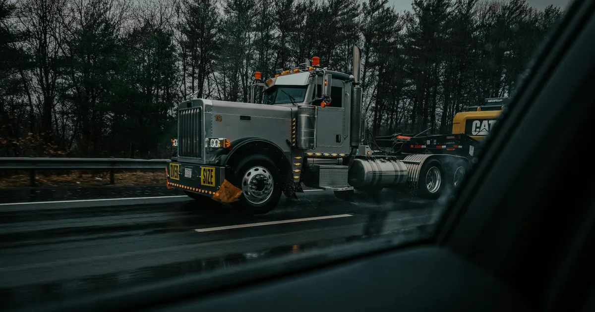 A large truck with an oversize load on a rainy highway, captured through a car window with dark, moody tones.