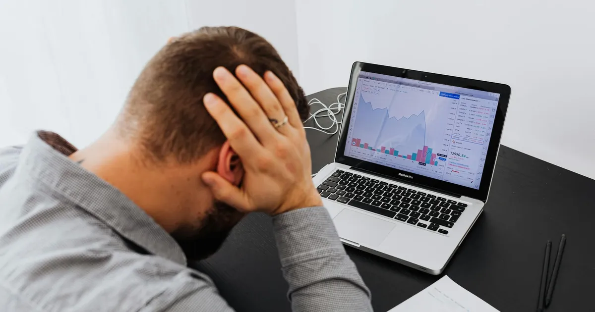 Stressed man at desk looking at declining stock charts on laptop, indicating financial loss.