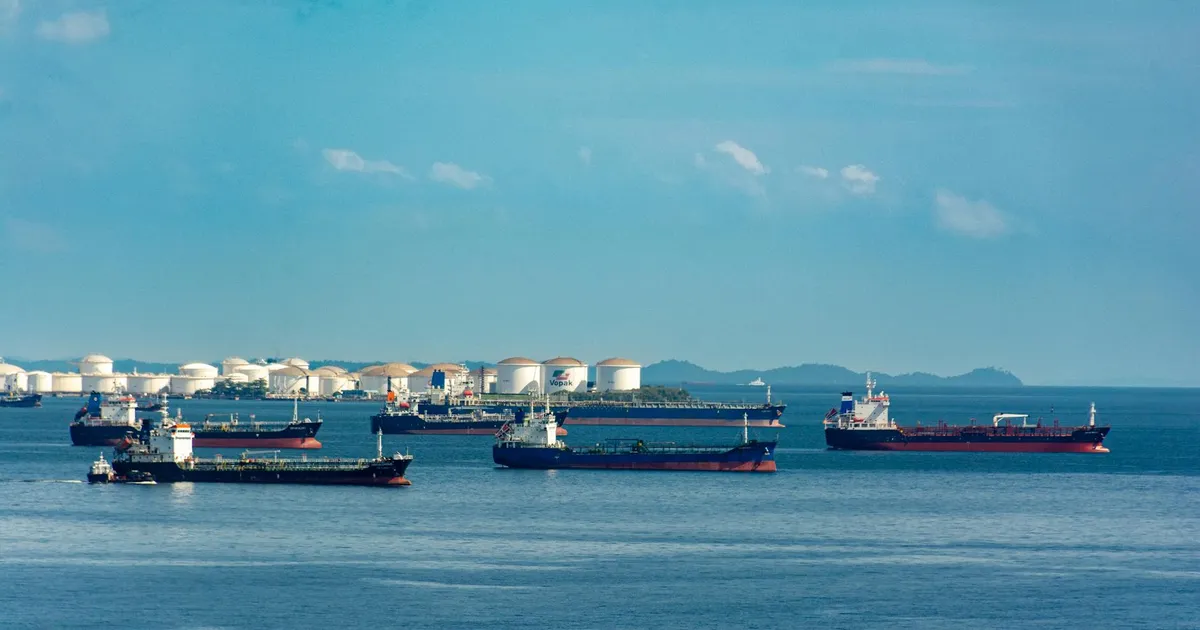 A fleet of cargo ships docked near oil storage tanks along a serene coastline with a clear blue sky above.